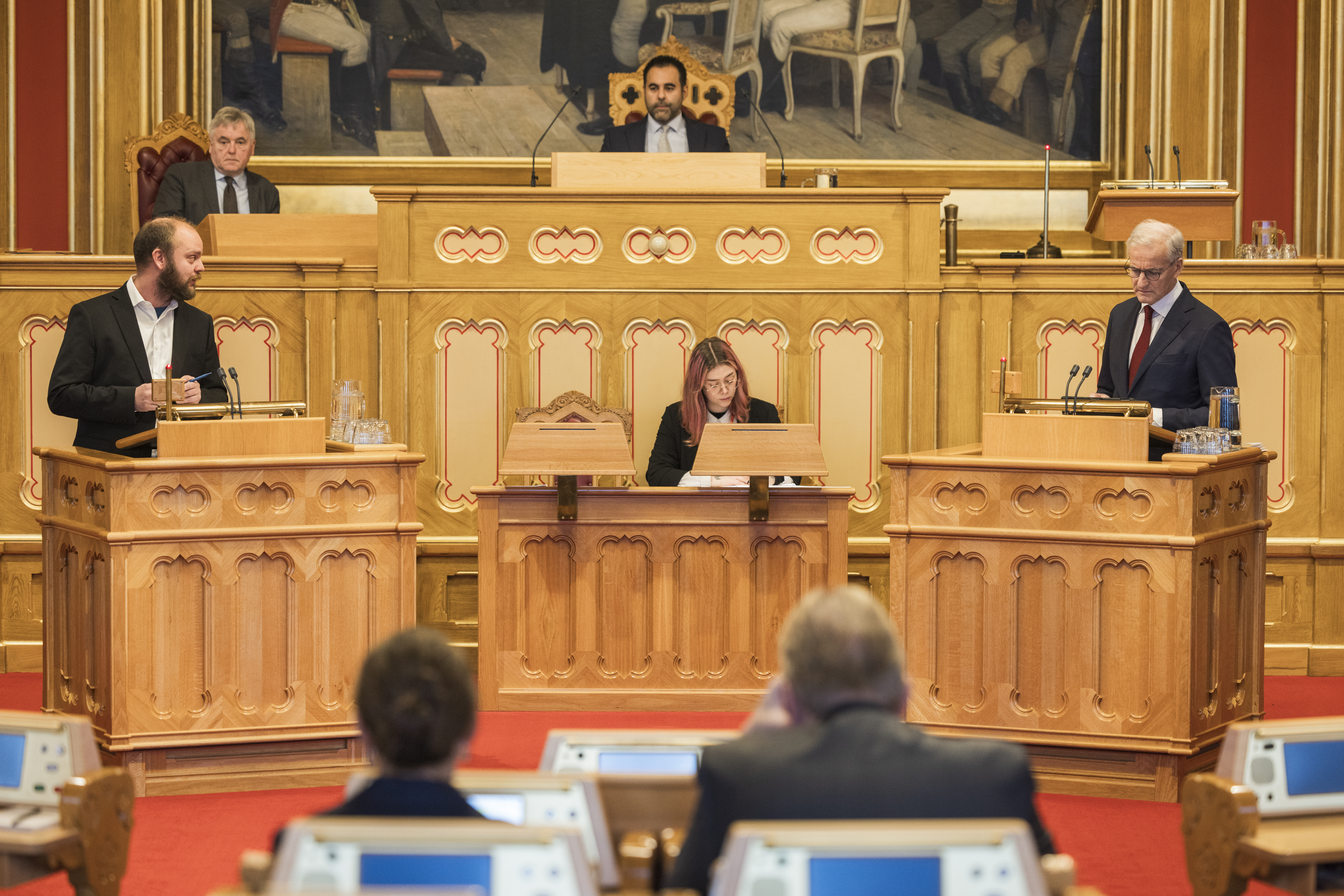 To personer står på hver sin talerstol. I midten sitter presidenten på podiet, og nedenfor sitter en person og noterer.