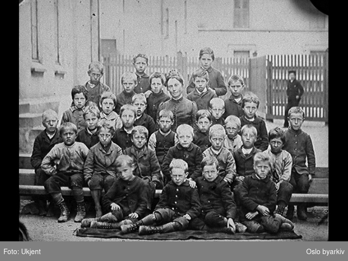 Svart hvitt bilde av en skoleklasse med gutter og en kvinnelig lærer i midten.