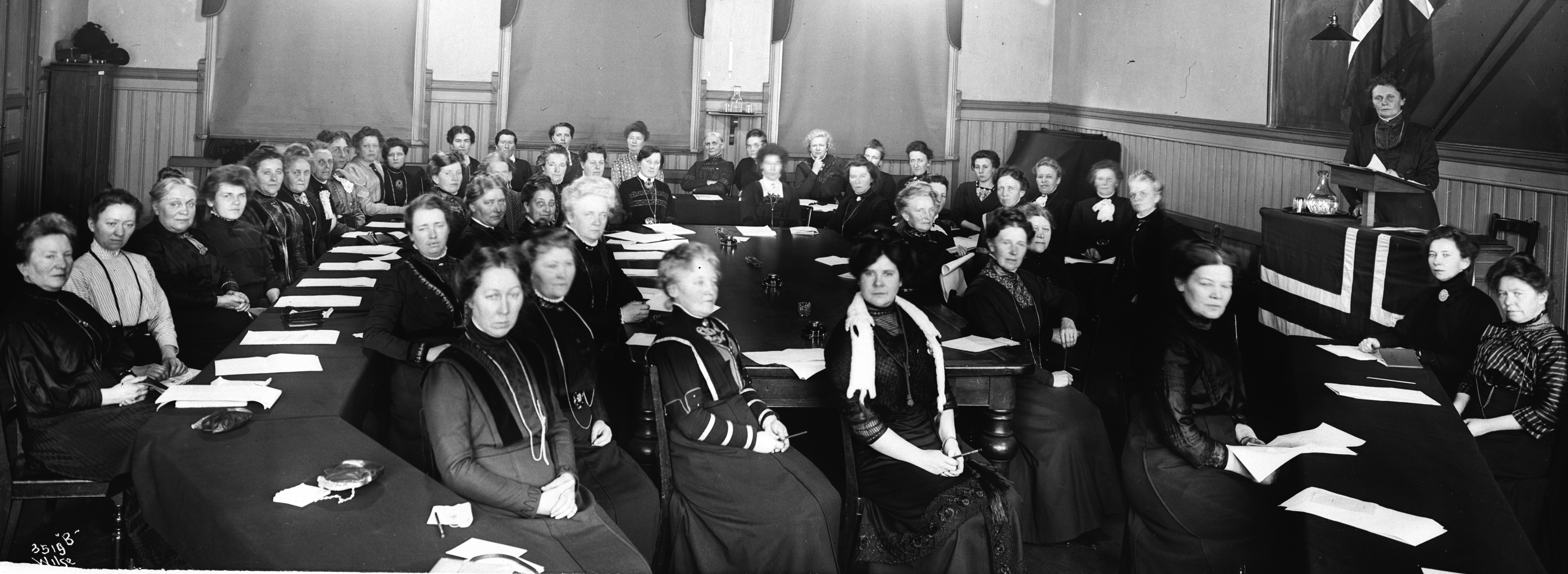 Svart hvitt bilde av mange kvinner som sitter i et rom. En kvinne står ved talerstolen øverst i rommet.