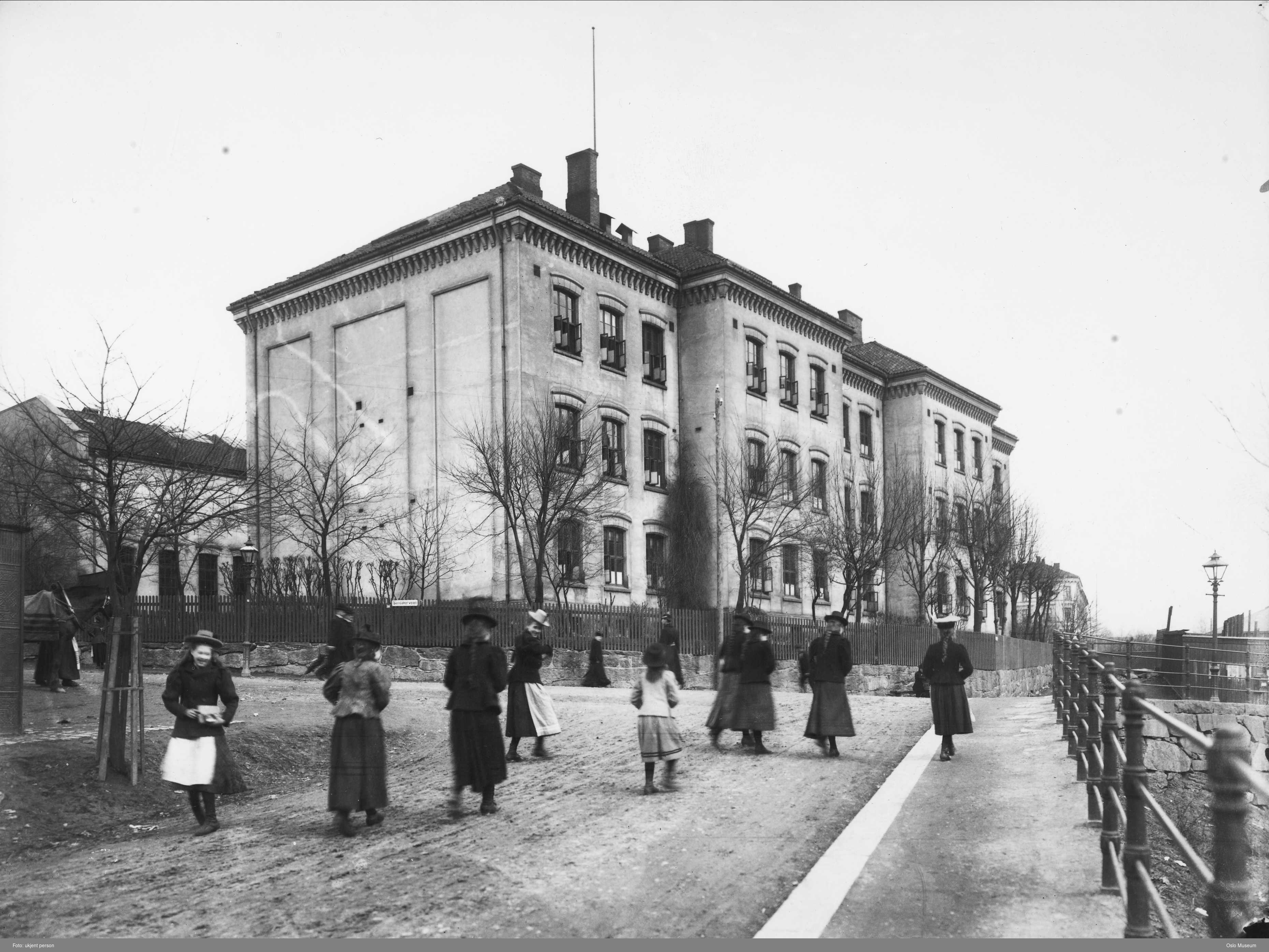 Svart hvitt bilde av en skole i mur, med mennesker som går forbi. 
