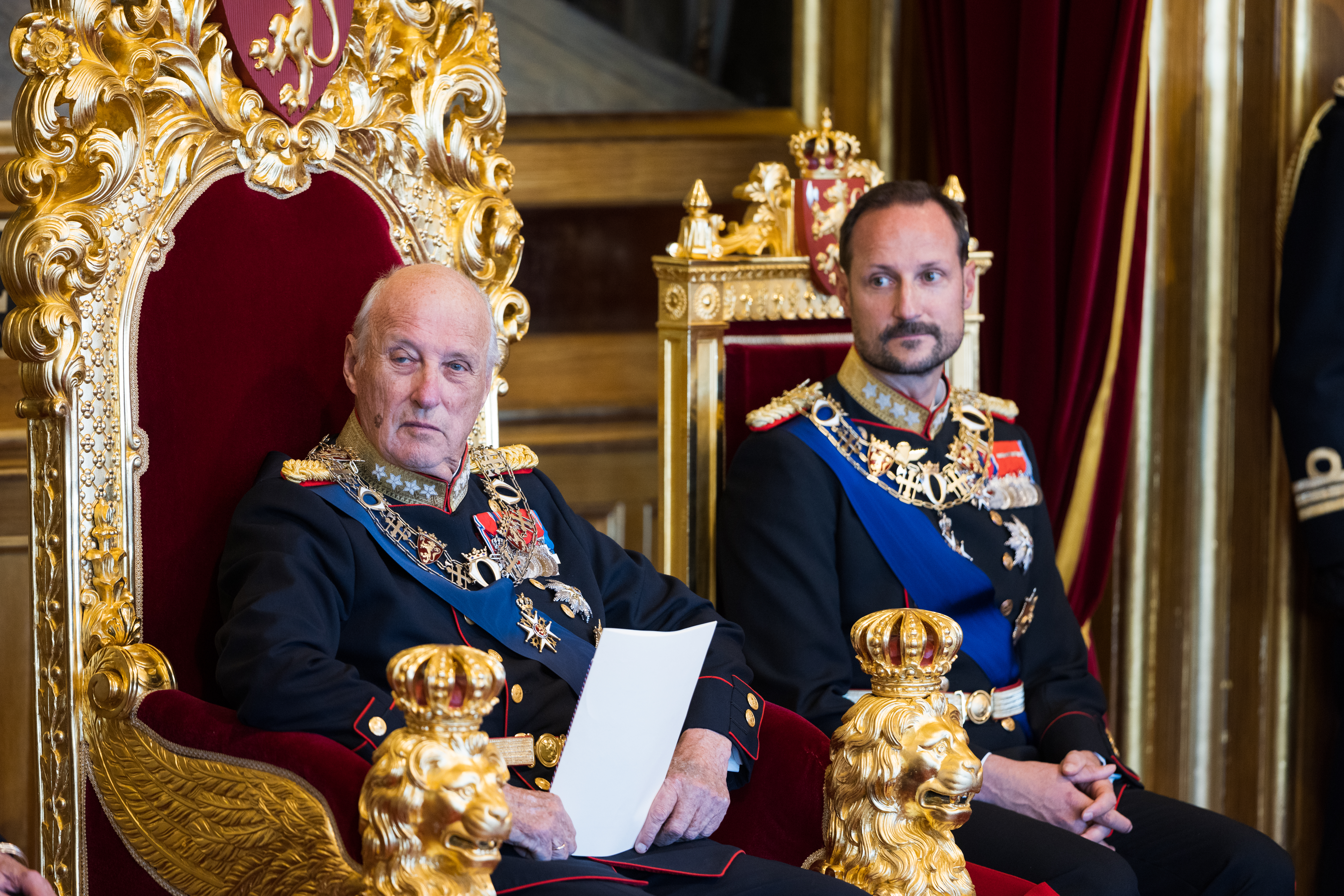 Kongen og kronprinsen sitter i hver sin gullforgylte stol. Kongen holder et ark i hånden.