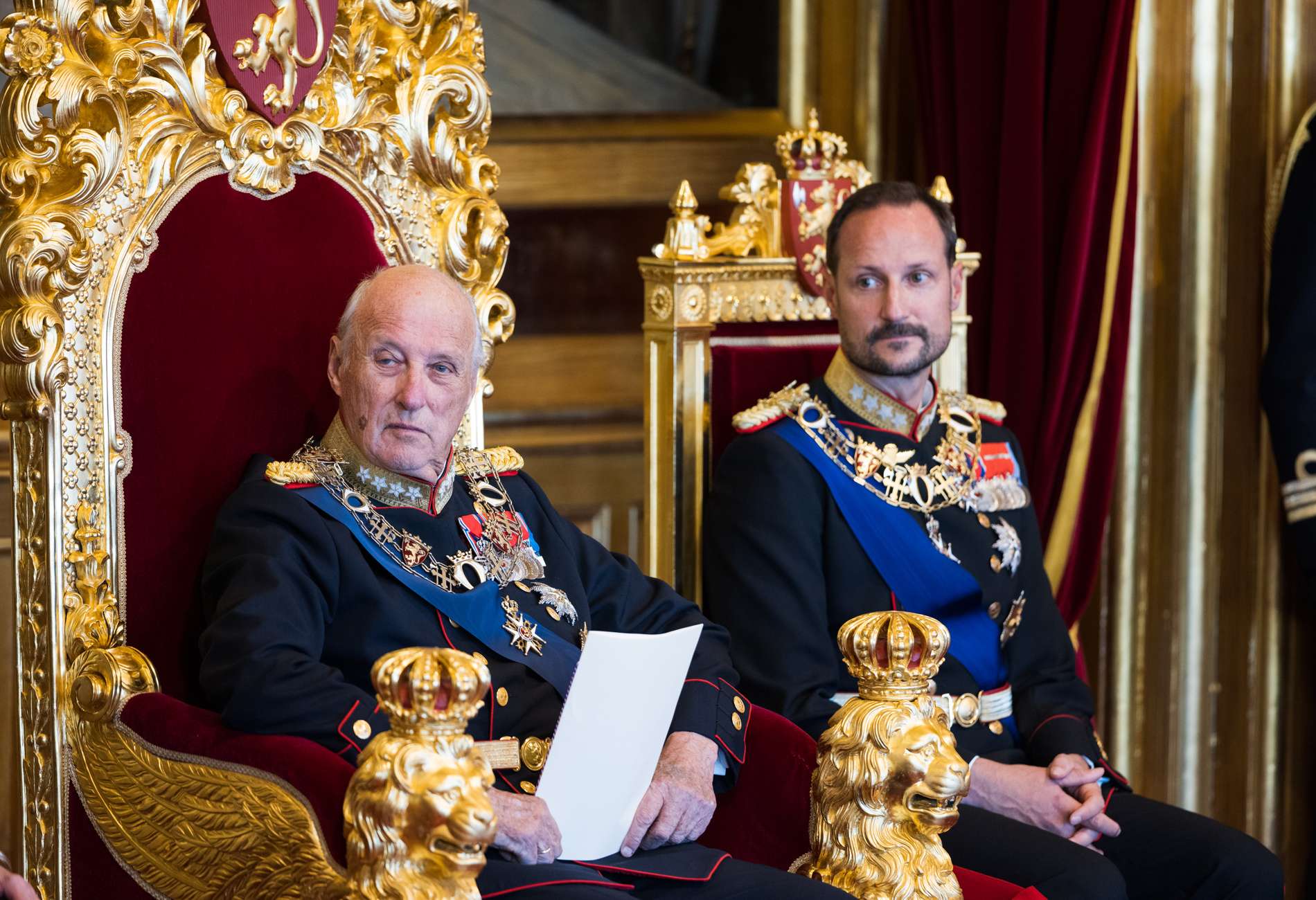 Kongen og kronprinsen sitter i hver sin gullforgylte stol. Kongen holder et ark i hånden.