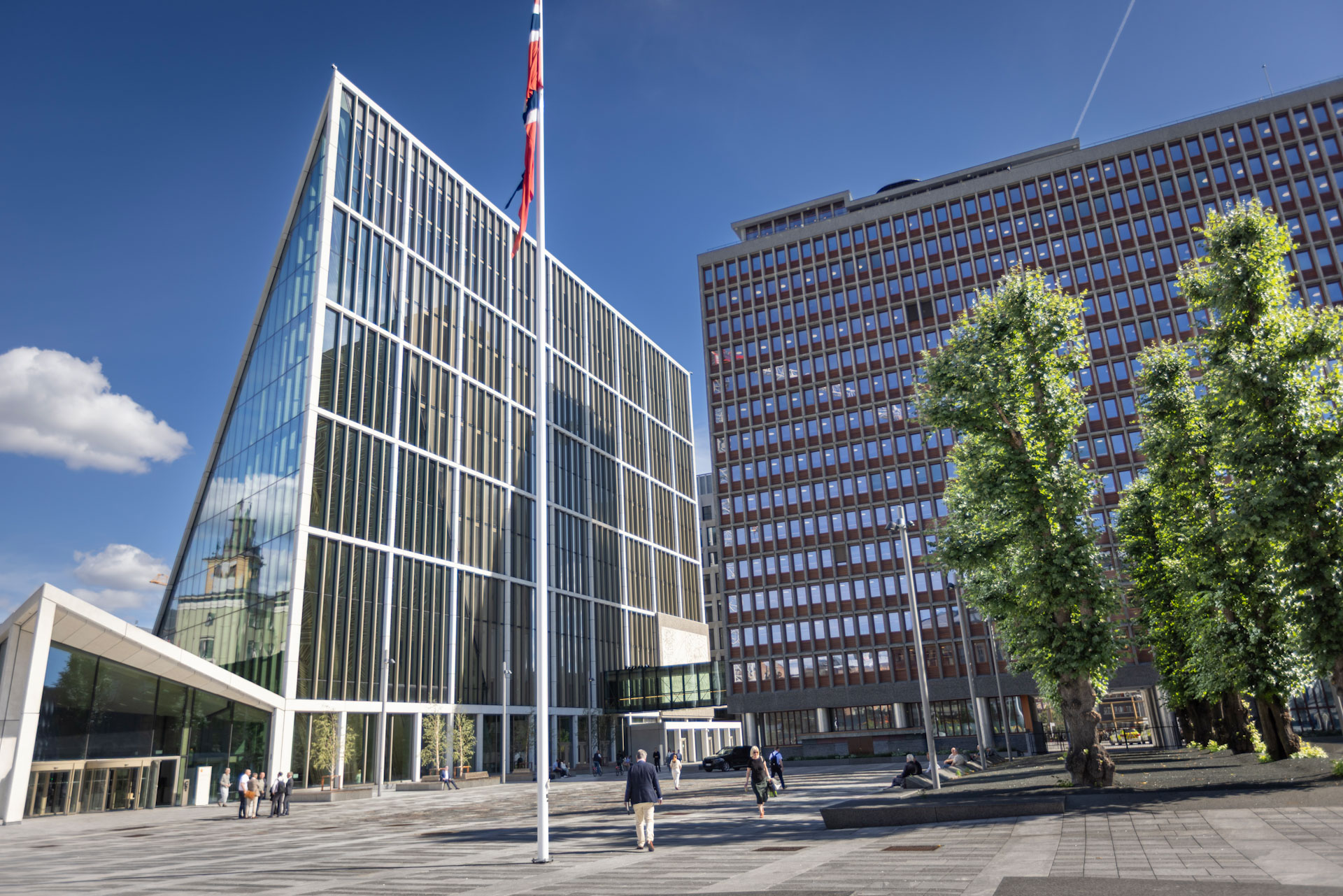 Store moderne bygninger rundt et åpent byrom med trær og mennesker.
