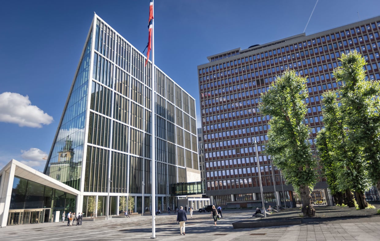 Store moderne bygninger rundt et åpent byrom med trær og mennesker.