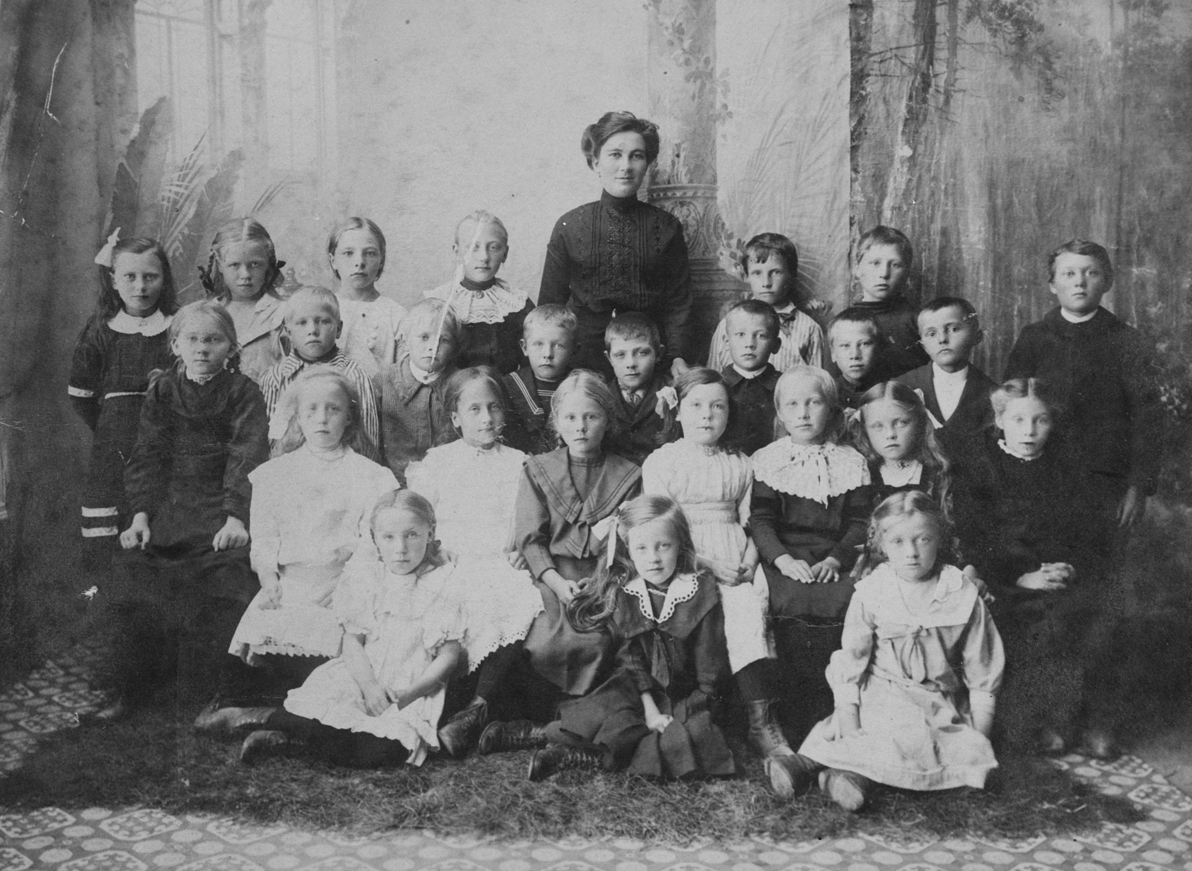 Svarthvitt bilde av en kvinnelig lærer med skoleklasse på 1800-tallet.