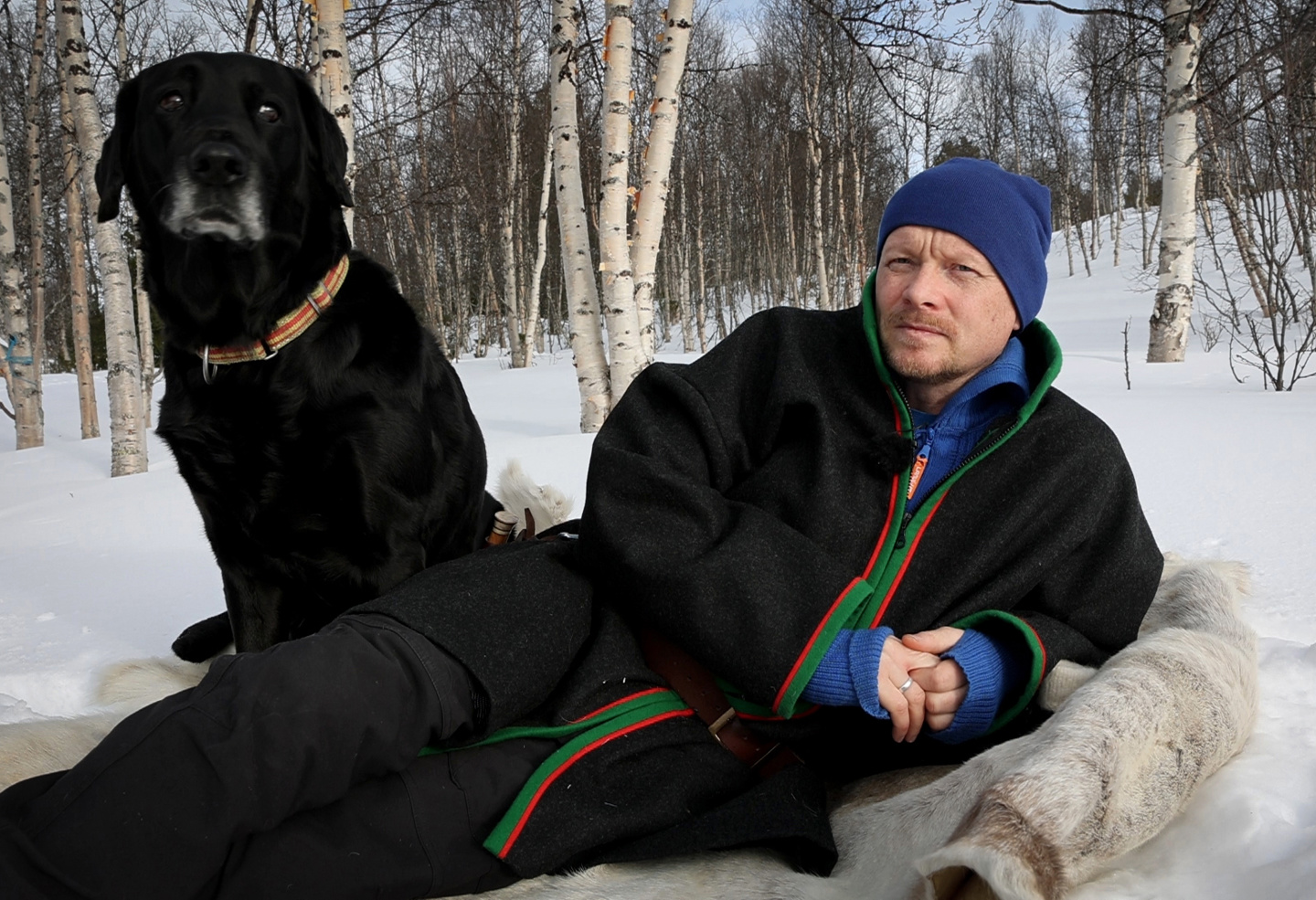 Mann ligger på skinnfell, hund sitter ved siden av.