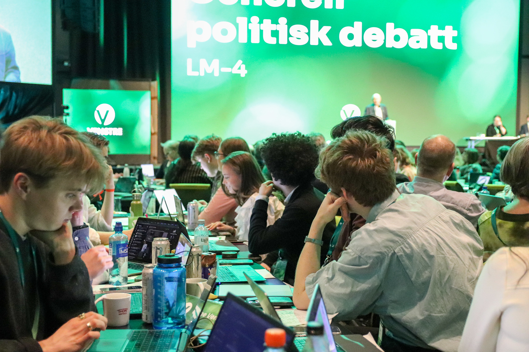 Mange mennesker sitter i en stor sal og jobber på pc. I bakgrunnen ses en scene med en person som snakker og partiet Venstres logo.