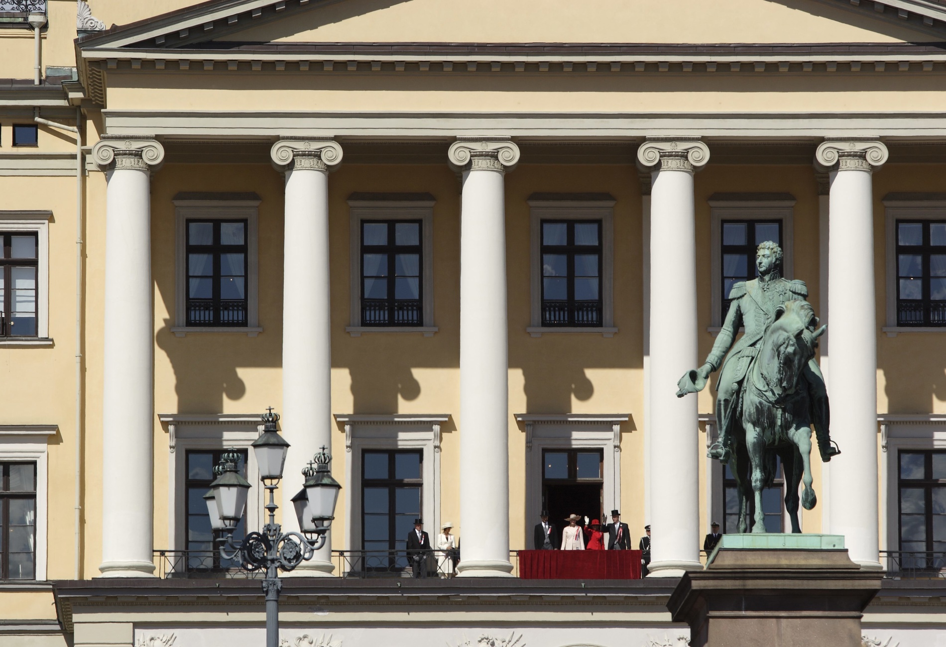 Kongefamilien på slottsbalkongen med statuen av Karl Johan i forgrunn.