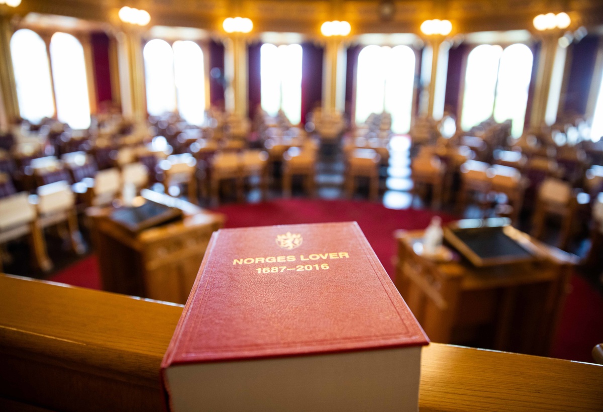 En lovbok ligger på en pult, stortingssalen er synlig i bakgrunn.