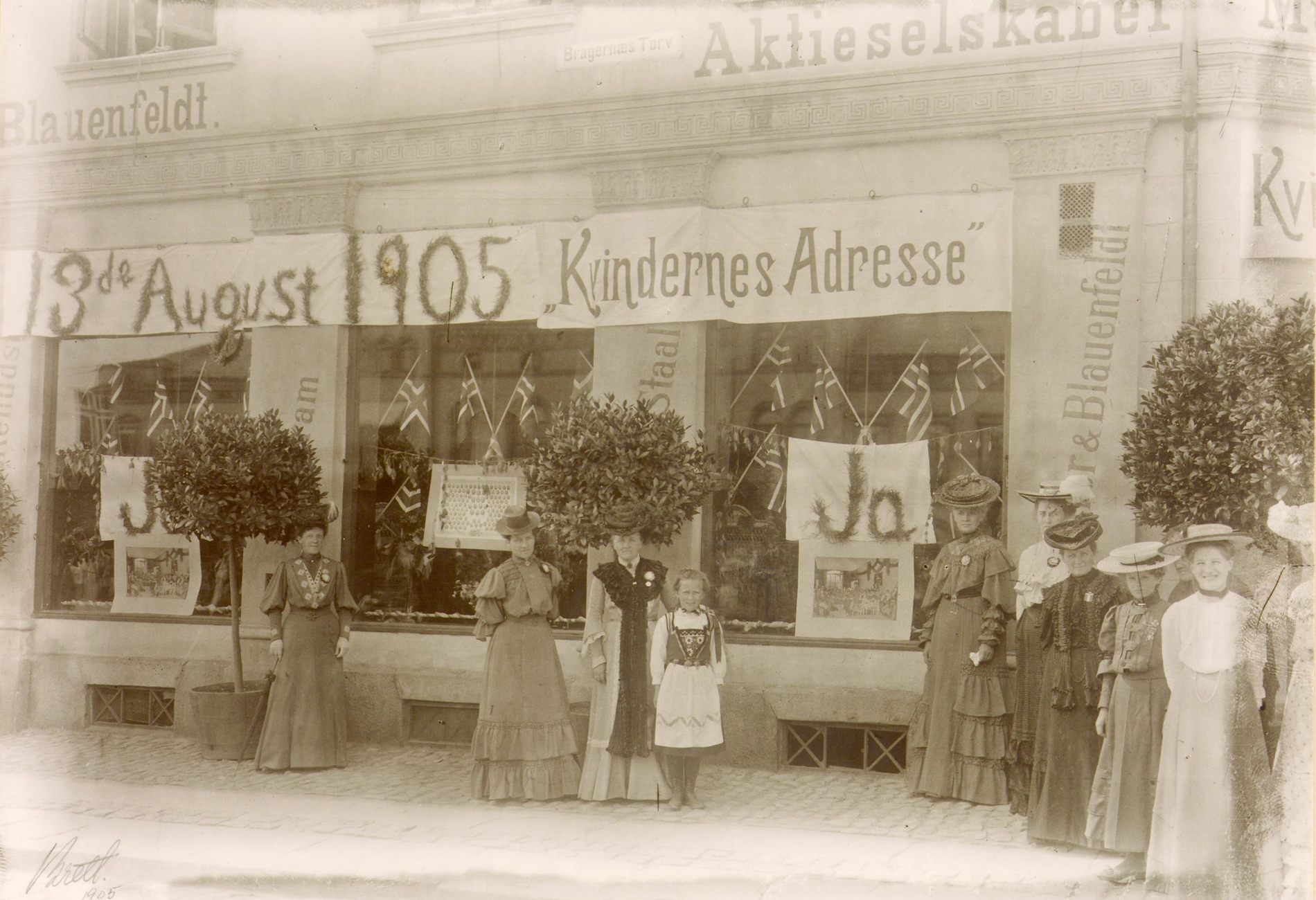 Svart hvitt fotografi av kvinner som står foran en butikk med et banner i bakgrunnen.