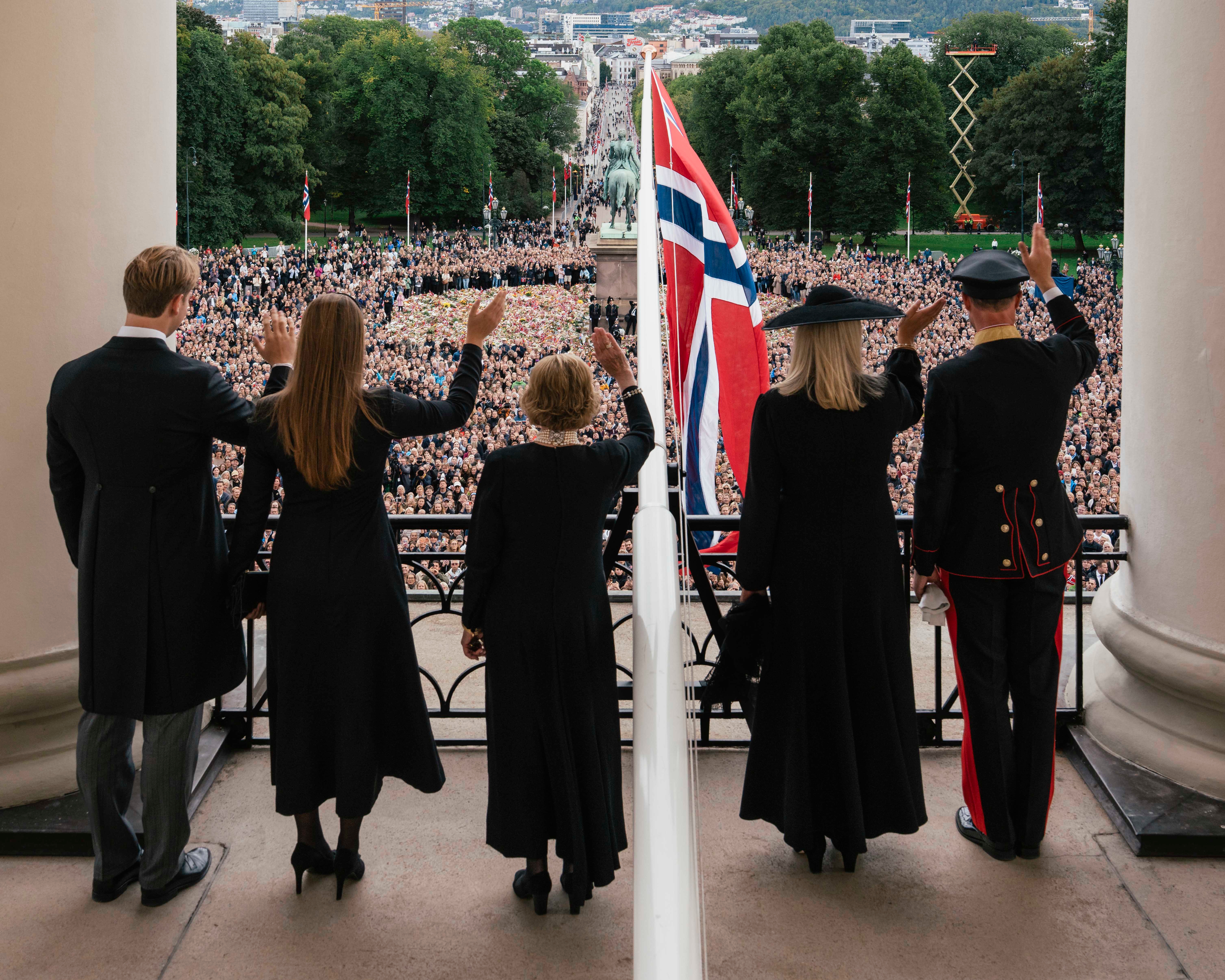 Kongefamilien og dronning Sonja står på slottsbalkongen. Det norske flagget er heist fra balkongen og de kongelige vinker til folkehavet på slottsplassen.