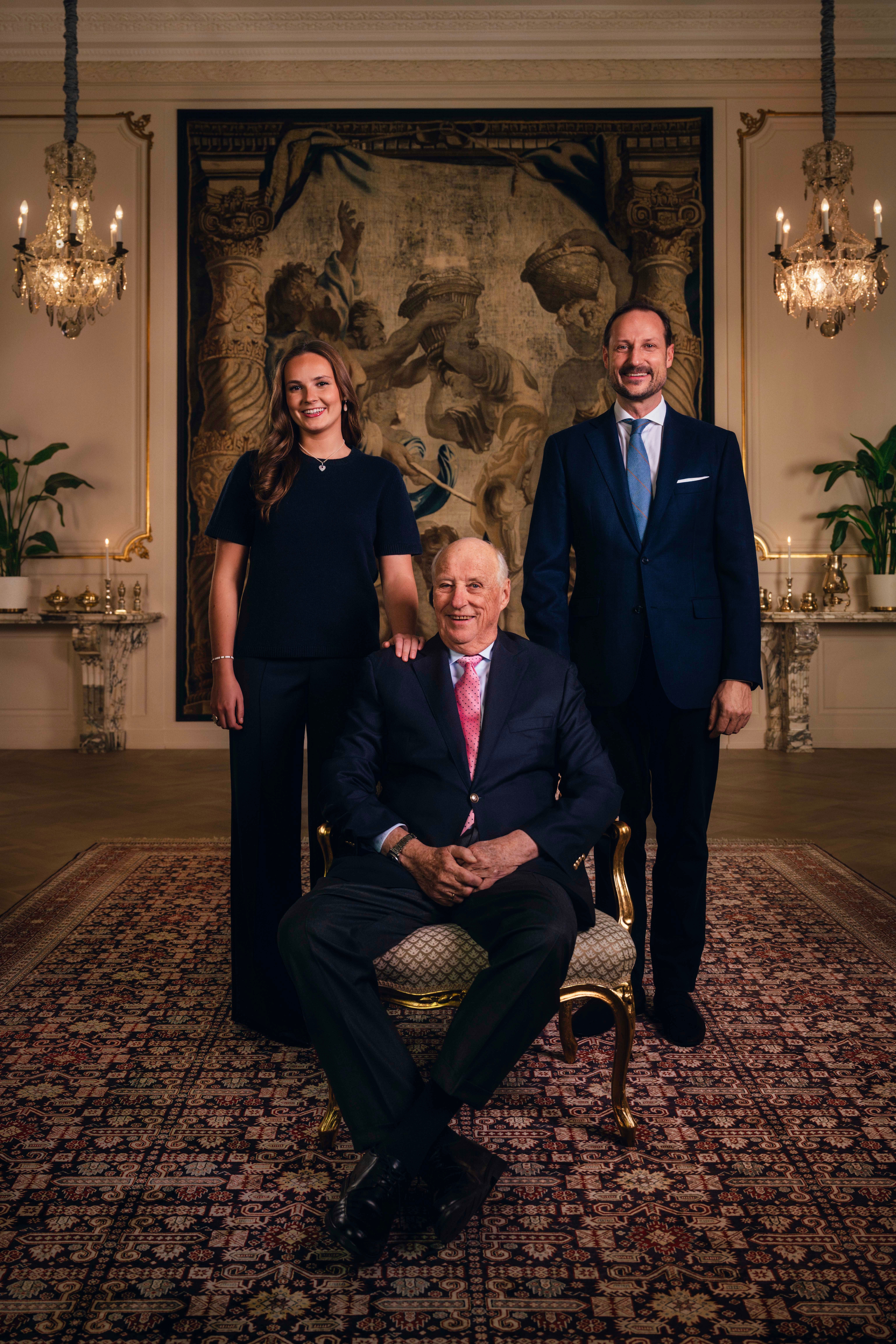 Kong Harald sitter på en stol. På kongens venstre side står prinsesse Ingrid Alexandra. På kongens høyre side står kronprins Haakon. 