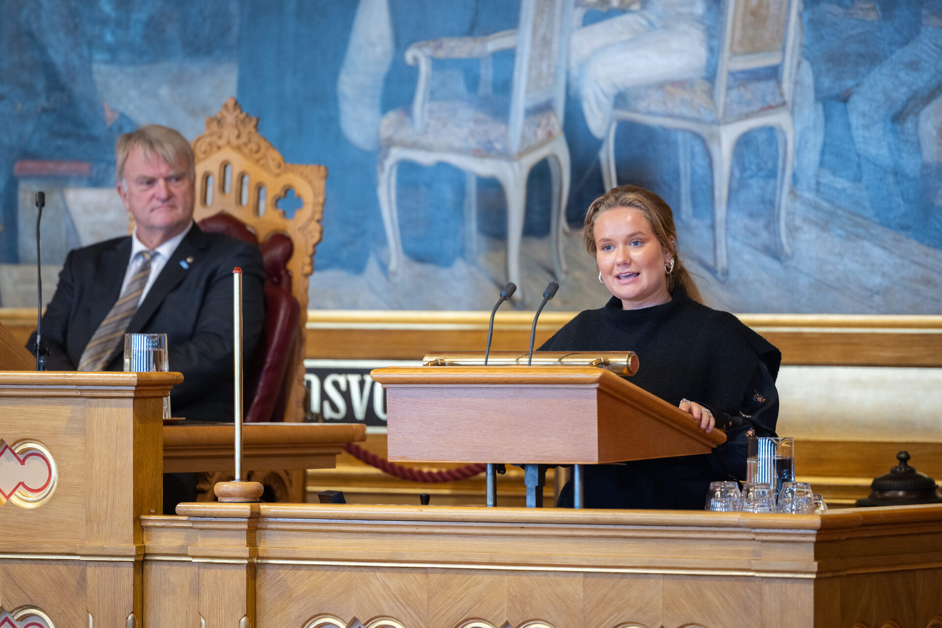 En ung kvinne står på talerstolen i stortingssalen. Stortingspresidenten sitter ved siden av.