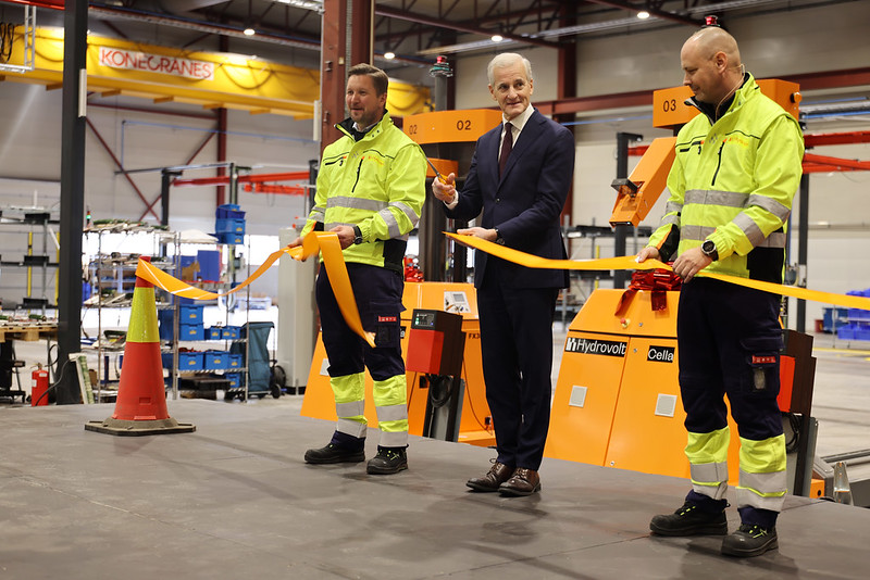 Statsminister klipper en gul snor på en fabrikk. To arbeidere holder snoren.