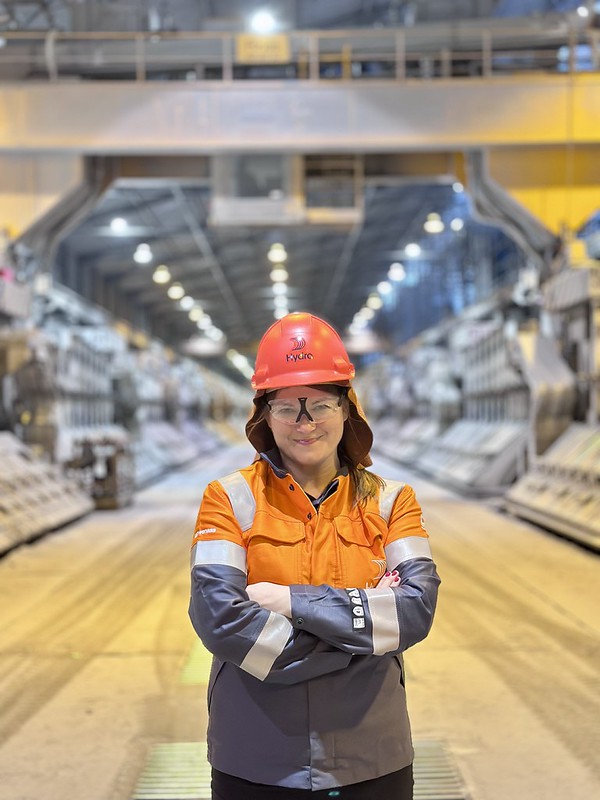 En kvinne i oransje jakke og oransje hjelm med skjerm, står med armene i kors. i bakgrunnen er det et fabrikklokale.