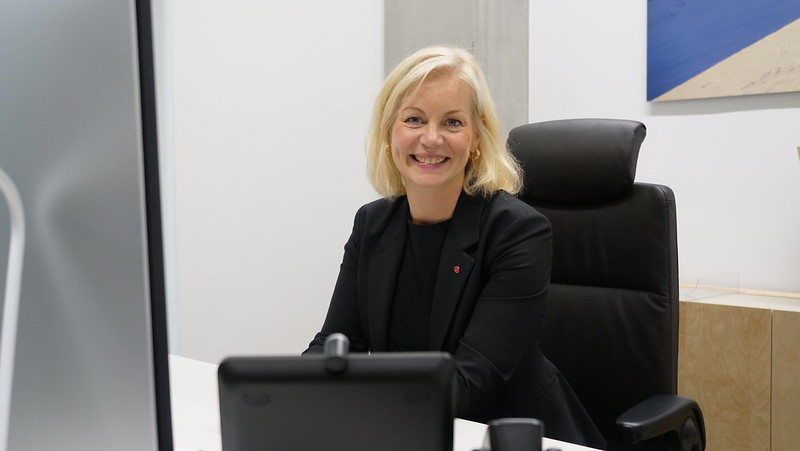 En kvinne sitter ved en pult og smiler til kamera.