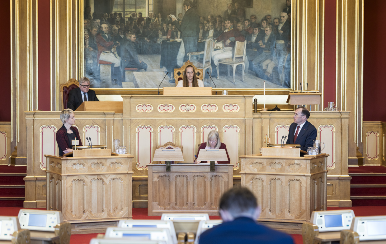 Stortingssalen. To politikere står på hver sin talerstol. Stortingspresidenten sitter i midten under Eidsvold 1814-maleriet.