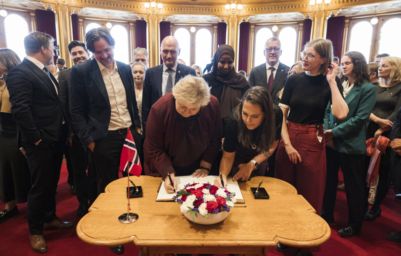 Mange stortingsrepresentanter står rundt et bord og venter på å signere.