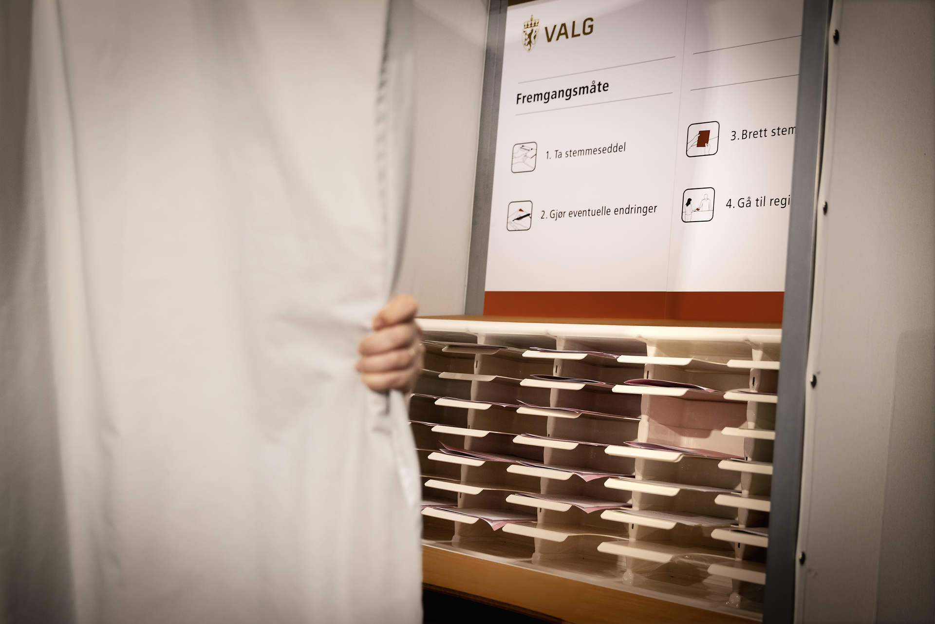 En halvåpen gardin med valgavlukke bak. Stemmesedler ligger i hyller.