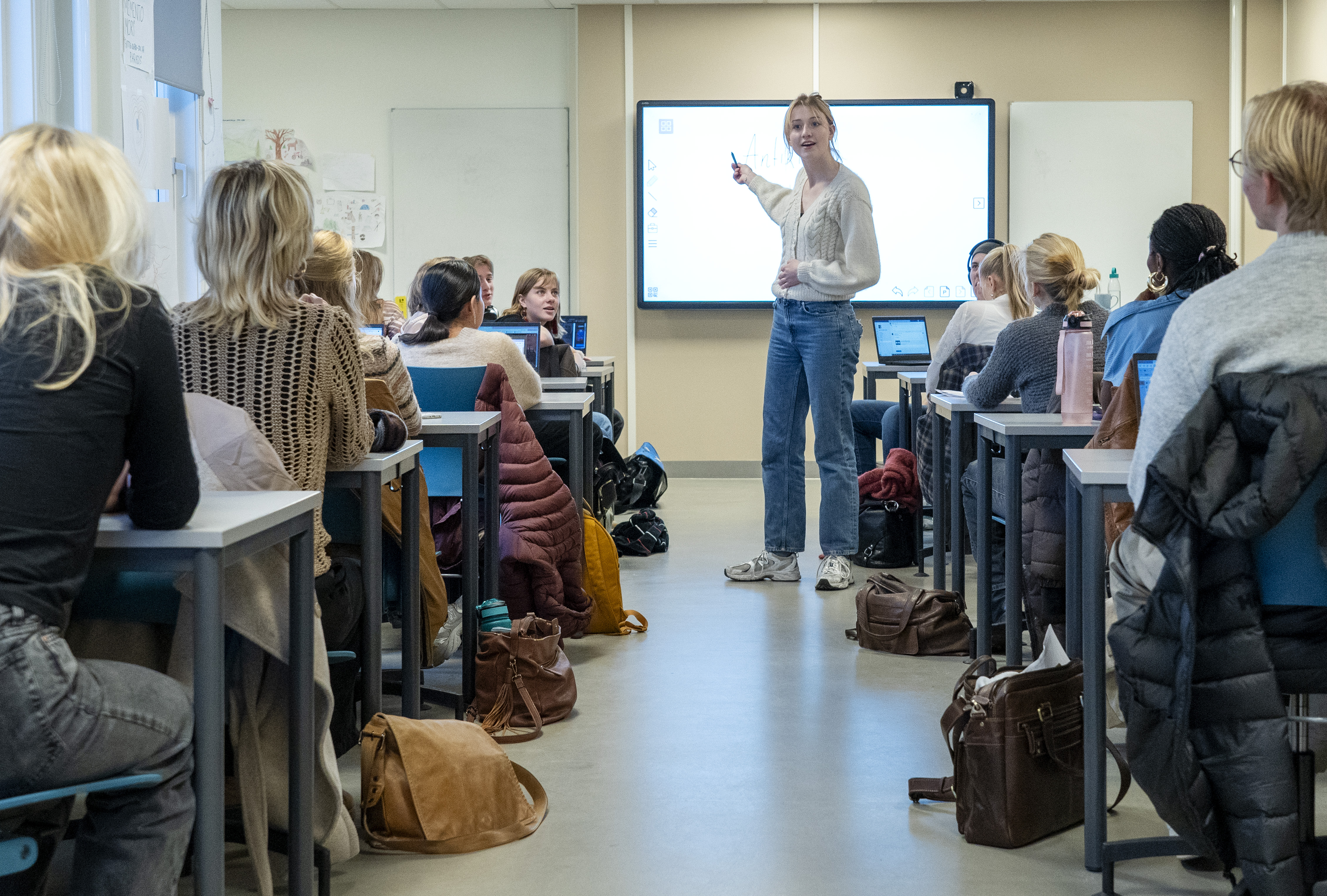 Ung kvinnelig lærer ser ut mot elever i klasserom mens hun peker på skjermen bak seg