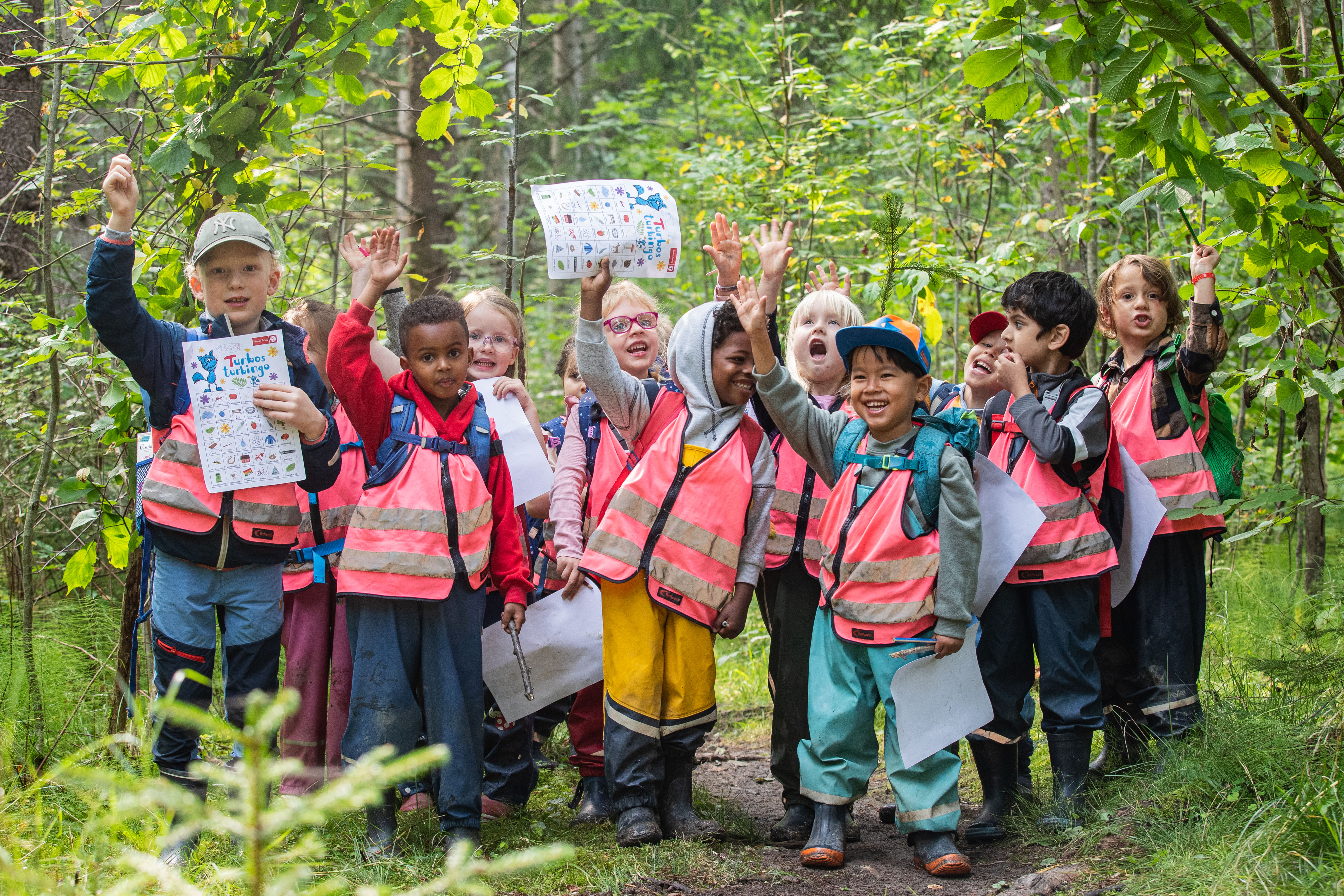 Barnehagebarn på tur i skogen. Barna poserer for bildet smilende med hendene i været.