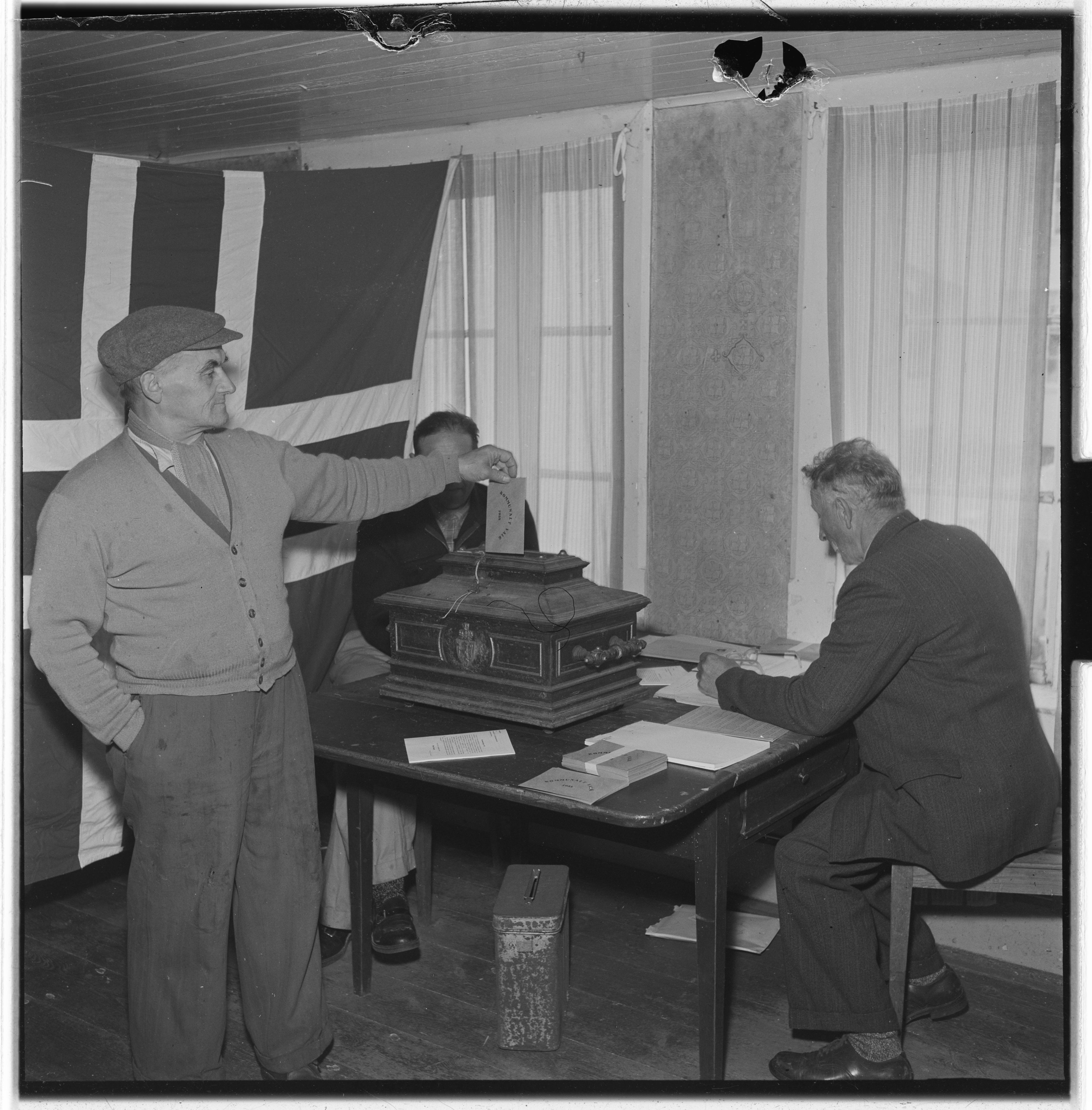 Svart hvitt bilde. En eldre mann med hatt legger en stemmeseddel i en valgurne. Det er et stort norsk flagg i bakgrunnen. Ved valgurnen sitter to menn.