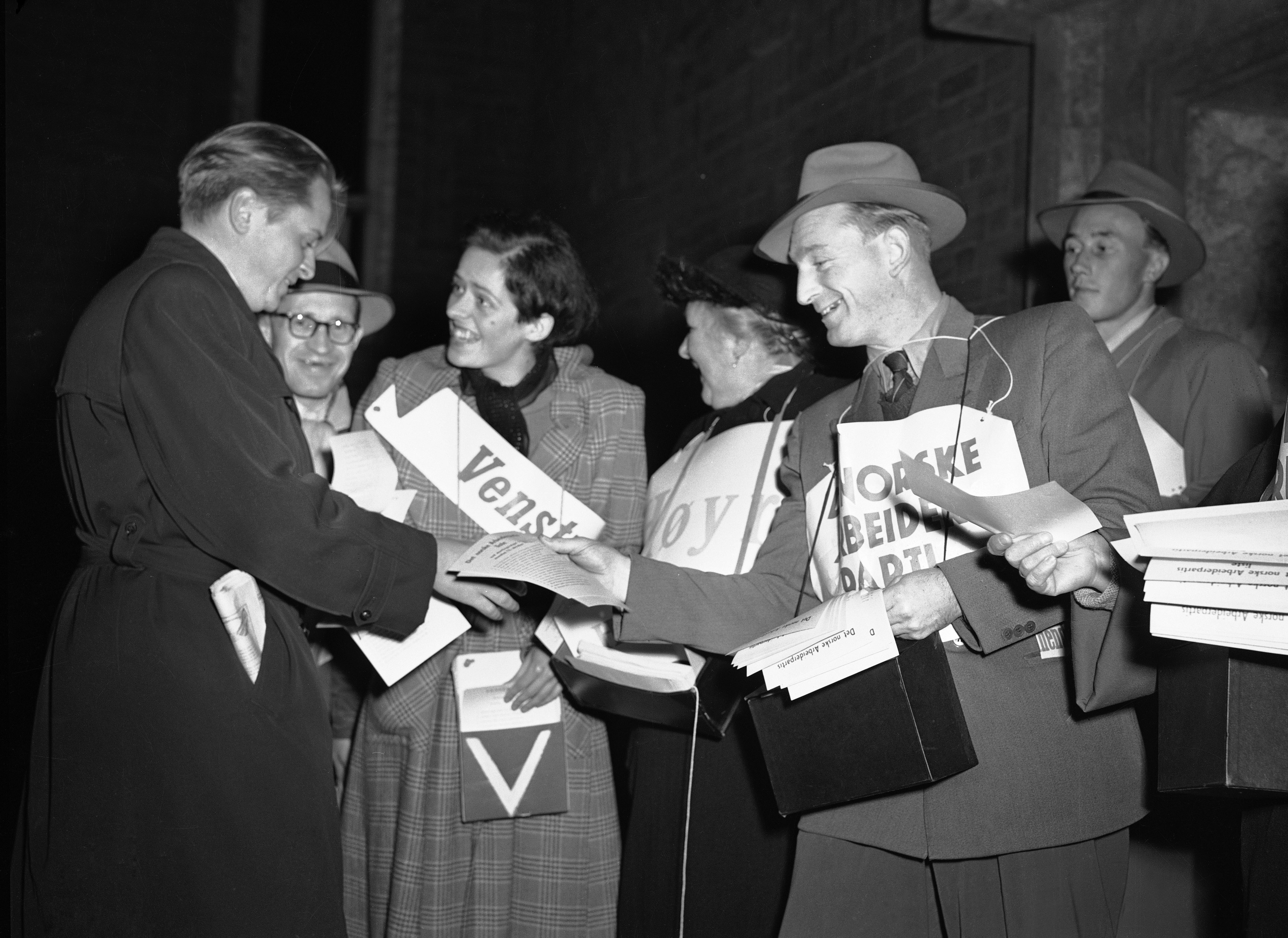 Svart hvitt bilde. Mennesker fra forskjellige partier deler ut løpesedler til en mann. 