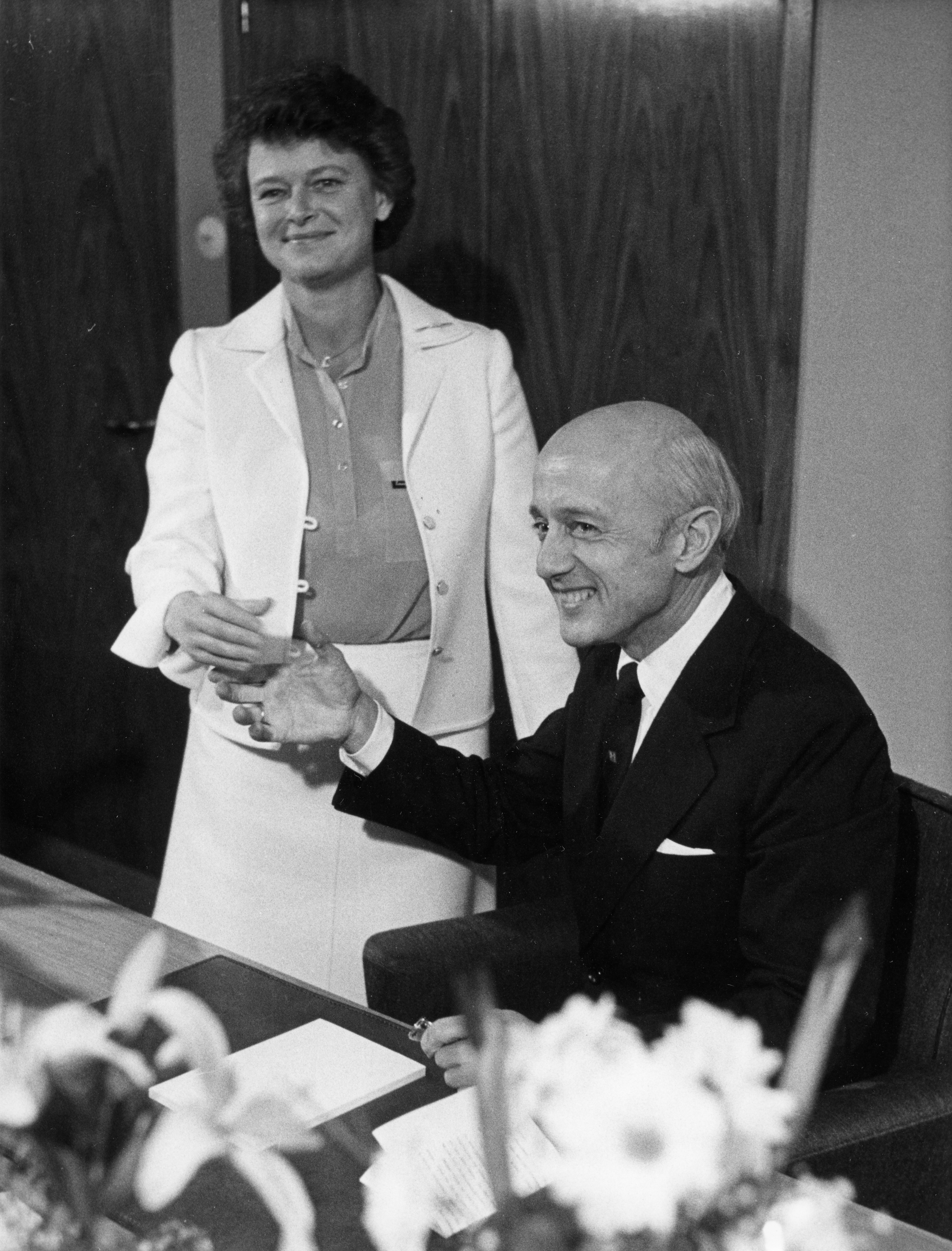 Svart hvitt bilde. Kåre Willoch sitter bak en pult. Gro Harlem Brundtland står ved siden av og gir han en nøkkel. Begge smiler.