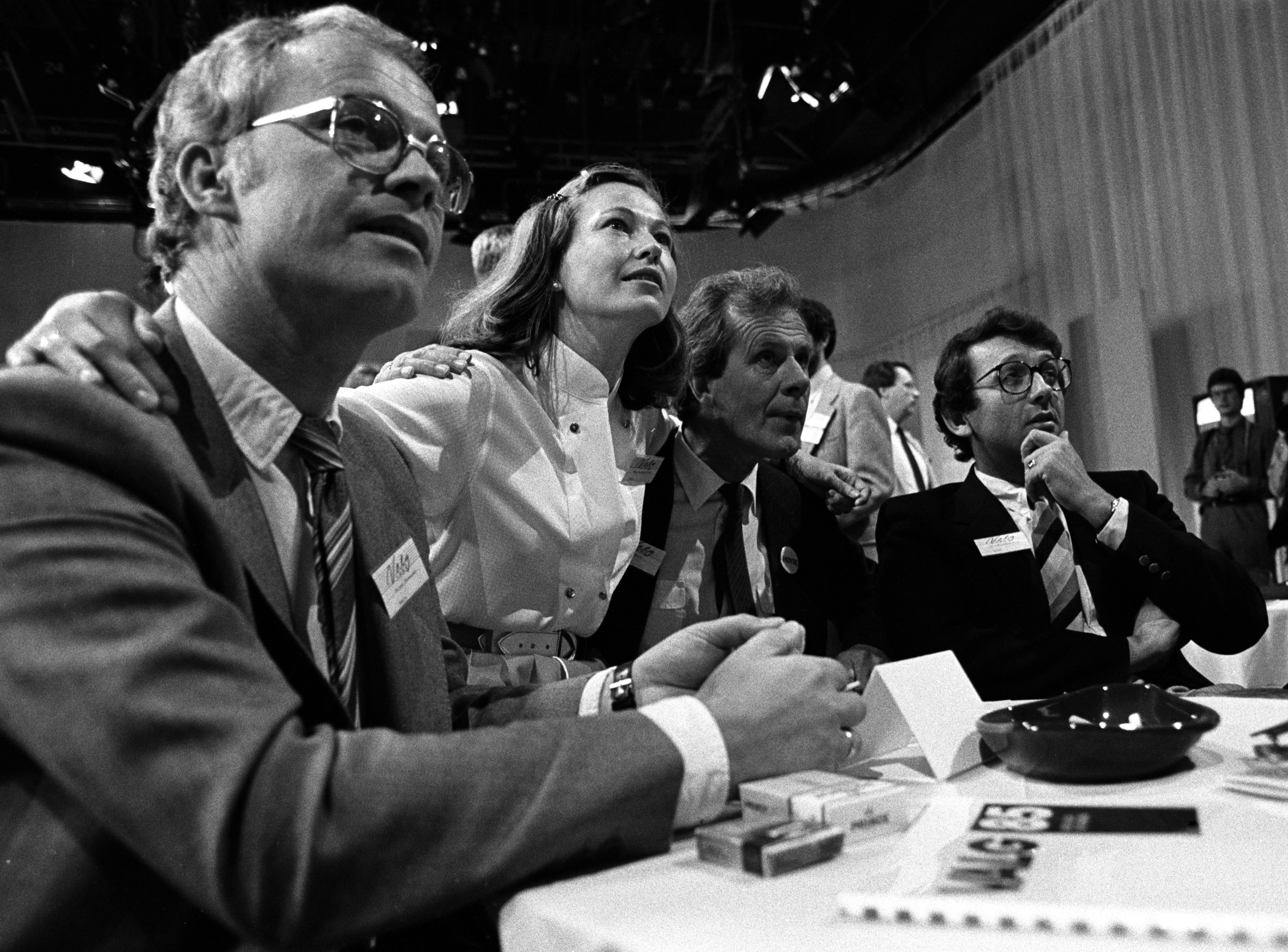 Svart hvitt bilde. Fire mennesker sitter rundt et bord og holder hverandre om skuldrene. De følger med på noe, antagelig en skjerm.