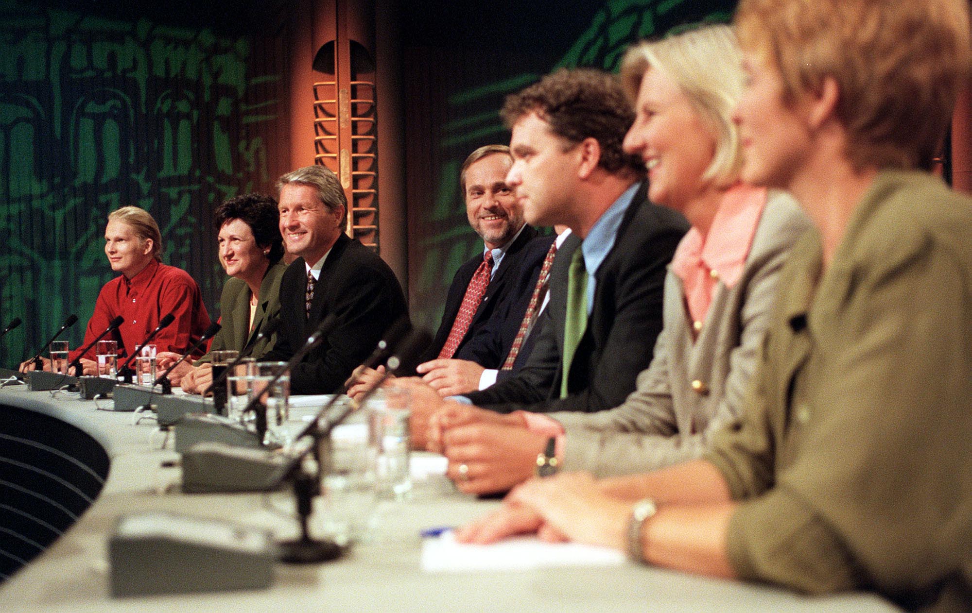 Syv politikere sitter i et debattpanel og smiler.