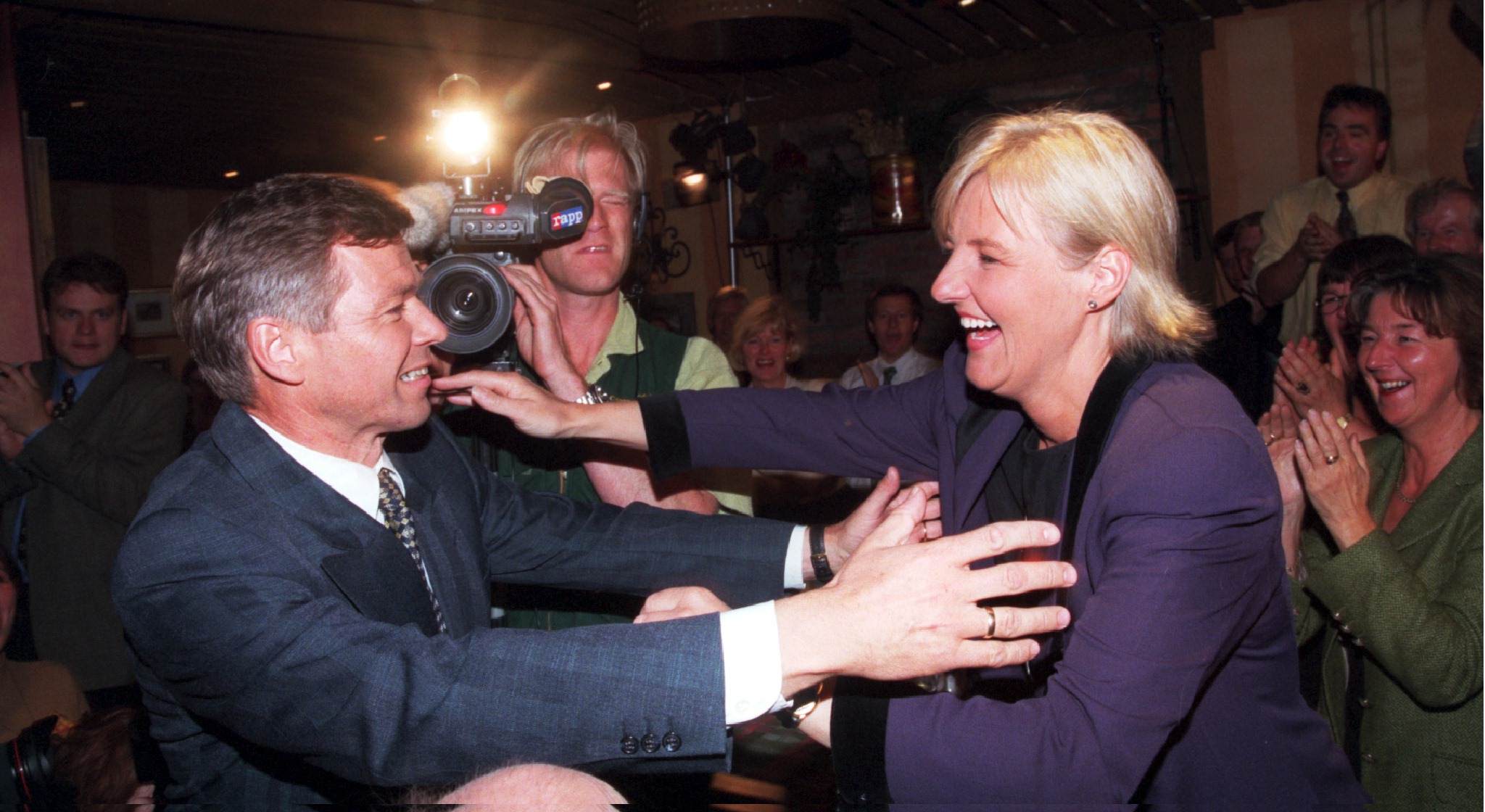 En mann og en dame er i ferd med å klemme hverandre. Bak står det en fotograf og mange glade mennesker.