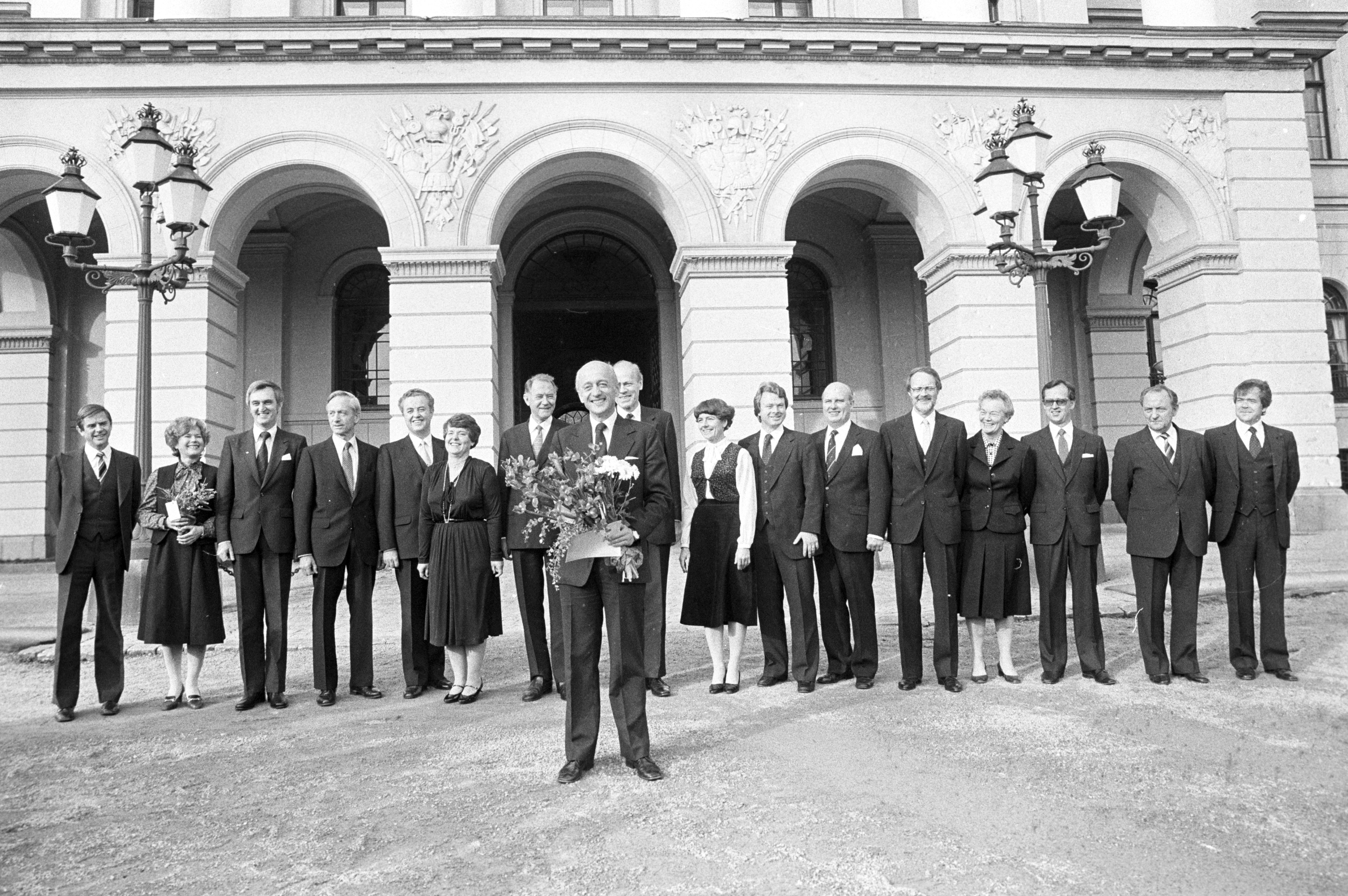 Svart hvitt bilde. En regjering står foran slottet i Oslo. Foran står statsminister Kåre Willoch med blomster i hendene. 
