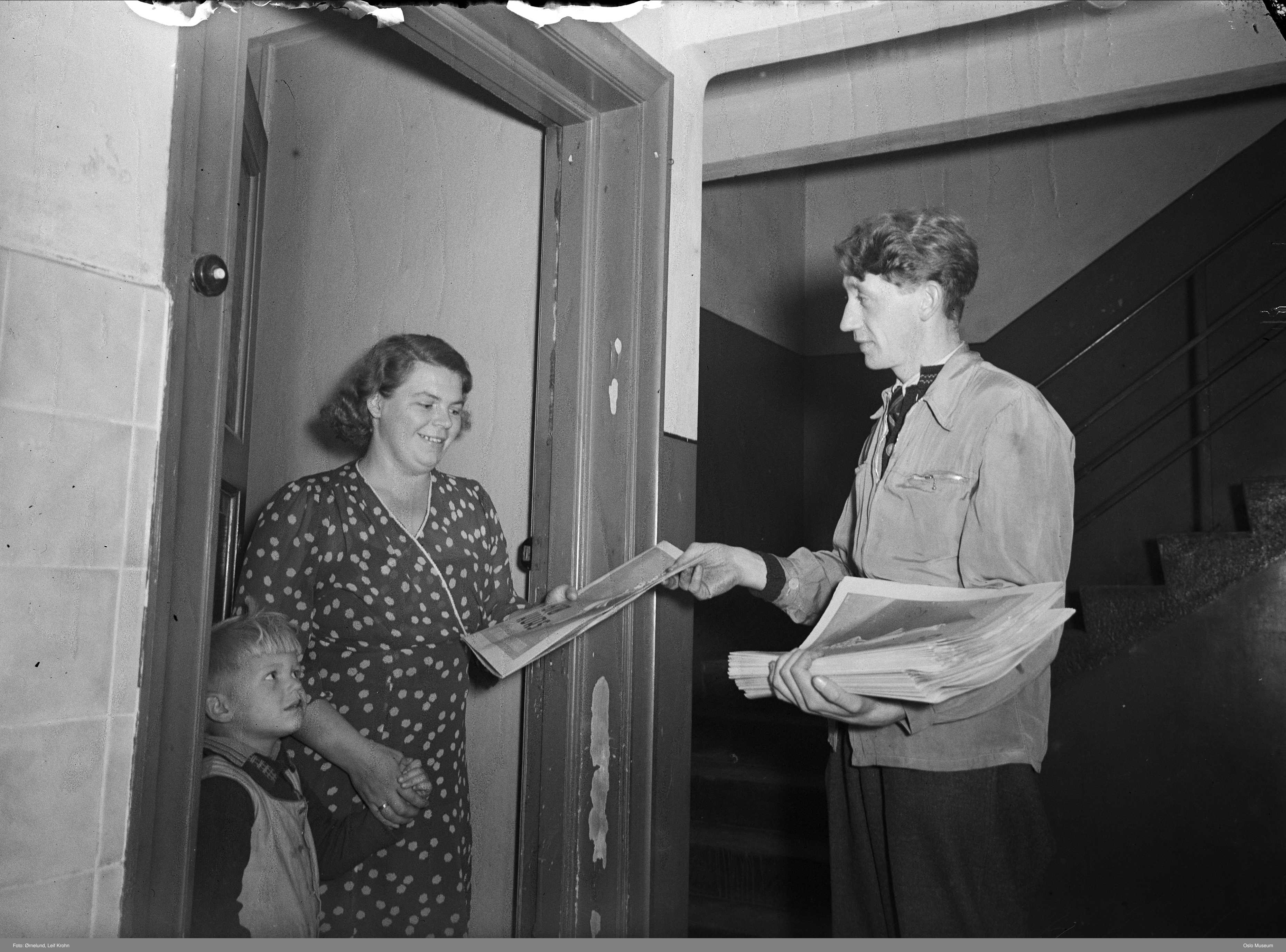 Svart hvitt bilde. En mann gir en avis til en kvinne og en barn som står i en åpen dør.