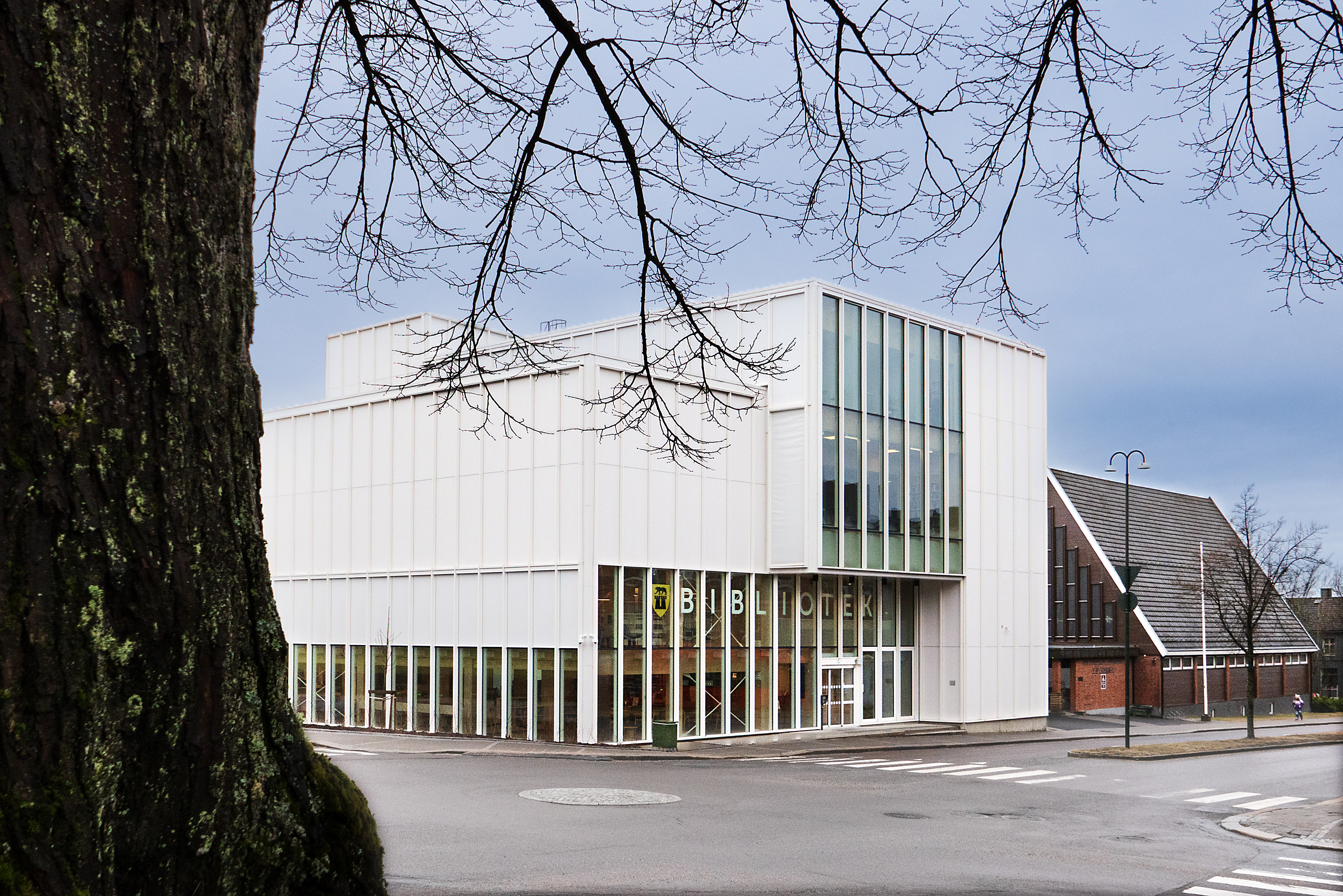 Hvit bygning med store vinduer i første og andre etasje. Gråblå himmel i bakgrunnen. Foto i farger av Sarpsborg bibliotek.